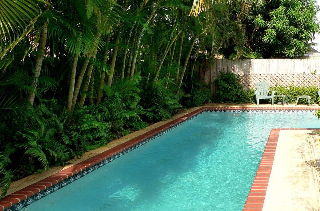 Older rectangular pool with brick coping and a stained, weathered concrete deck beside a wood fence
